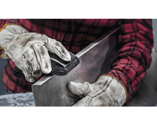 Eine Person reinigt ein Metallstück mit einer Schleifbürste, geschützt durch Arbeitshandschuhe und ein kariertes Hemd.