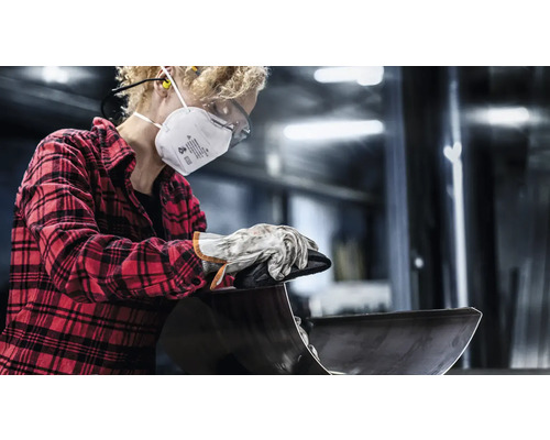 Eine Person mit Atemschutzmaske, Schutzbrille und Handschuhen bearbeitet ein Metallwerkstück.