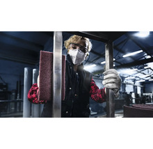 Eine Person mit Schutzbrille und Atemschutzmaske hält ein Schleifvlies in einer Werkstatt.