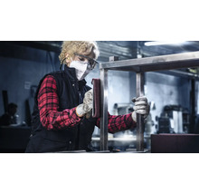 Eine Frau mit Schutzmaske und Schutzbrille schleift ein Metallgestell in einer Werkstatt.