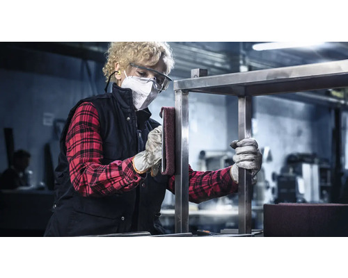 Eine Frau mit Schutzmaske und Schutzbrille schleift ein Metallgestell in einer Werkstatt.