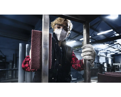 Frau mit Schutzbrille und Atemschutzmaske hält Schleifvlies zur Metallbearbeitung.
