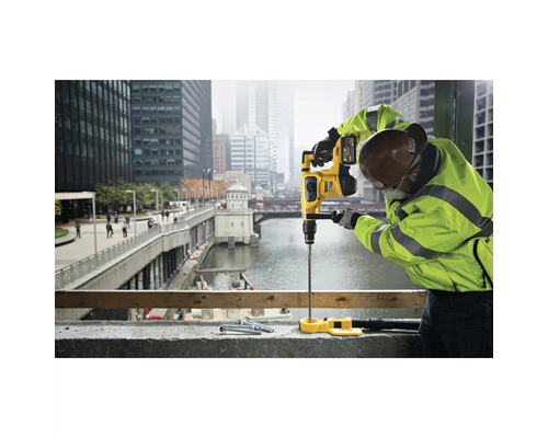 Ein Bauarbeiter bohrt mit einem Bohrhammer ein Loch in eine Betonoberfläche. Im Hintergrund befindet sich eine Stadt.