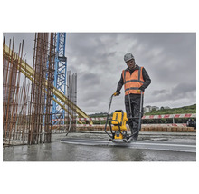 Ein Bauarbeiter verwendet eine Betonabziehmaschine auf einer Baustelle, im Hintergrund befinden sich Baustahl und ein Kran.