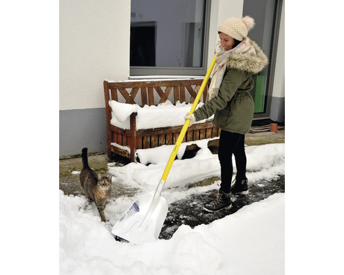 Frau schippt Schnee mit einer Schneeschaufel vor dem Haus, während eine Katze zusieht.