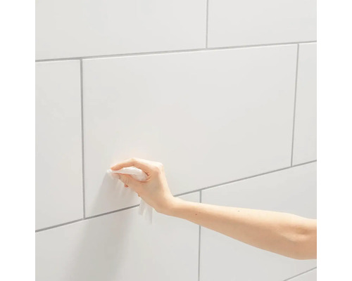 Eine Hand reinigt eine weiße geflieste Wand mit einem Tuch.