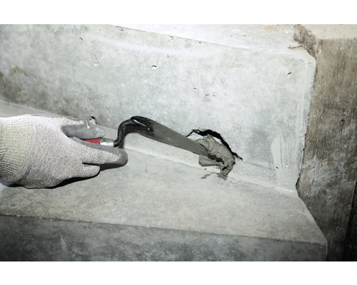 Eine Hand mit Schutzhandschuh verschließt ein Loch in einer Betontreppe mit einer Maurerkelle und Fugenmasse.