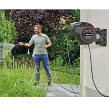 Ein Mann bewässert einen Garten mit einer automatischen Schlauchtrommel von Einhell an der Wand.