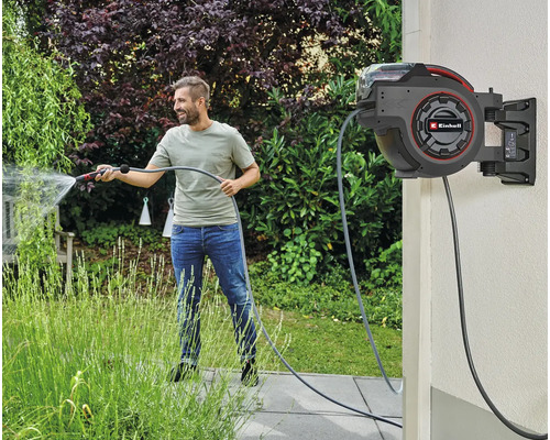 Ein Mann bewässert einen Garten mit einer automatischen Schlauchtrommel von Einhell an der Wand.