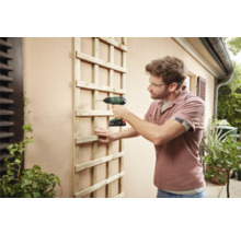 Ein Mann befestigt eine Holzspalierwand mit einem Akku-Bohrschrauber an einer Wand im Außenbereich.