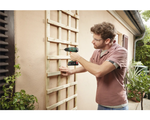 Ein Mann befestigt eine Holzspalierwand mit einem Akku-Bohrschrauber an einer Wand im Außenbereich.