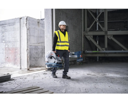 Ein Bauarbeiter mit Schutzhelm und Warnweste trägt eine Bosch Metallsäge auf einer Baustelle.