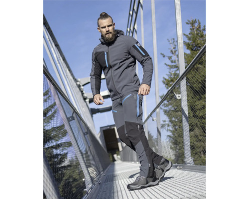 Mann in Outdoorjacke, Hose und Wanderschuhen auf einer Brücke.