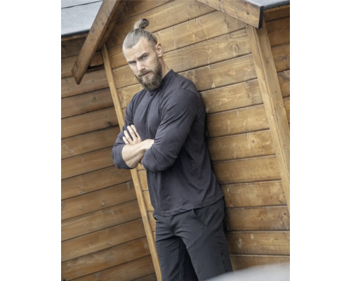 Mann mit Bart und Haarknoten lehnt an Holzhauswand.