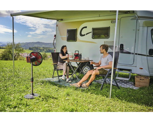 Szene mit Wohnmobil, Markise, Tisch, Stühlen und Personen auf einer Wiese.