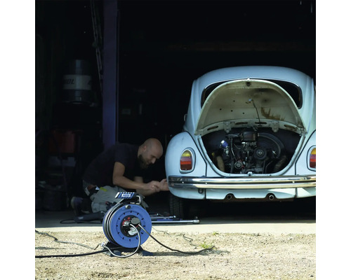 Eine Person repariert ein Auto mit geöffnetem Motorraum und einer Kabeltrommel im Vordergrund.