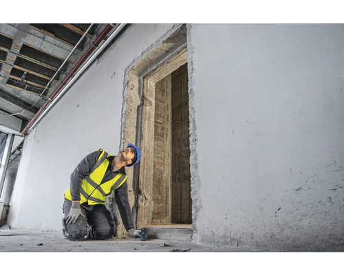 Ein Bauarbeiter mit Schutzhelm und Sicherheitsweste misst eine Türöffnung mit einem Laser Entfernungsmesser.