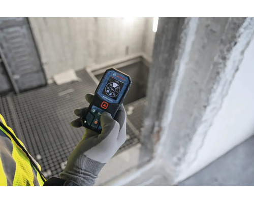 Hand mit Handschuh hält ein Bosch Entfernungsmessgerät in einem Rohbau.