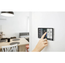 Aufnahme eines modernen Smart Home Touchpanels an der Wand, das von einer Hand bedient wird. Im Hintergrund ist ein heller Wohnbereich zu sehen.