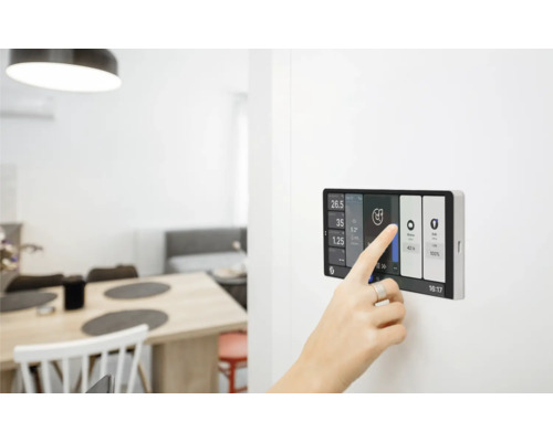 Aufnahme eines modernen Smart Home Touchpanels an der Wand, das von einer Hand bedient wird. Im Hintergrund ist ein heller Wohnbereich zu sehen.