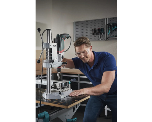 Mann mit Schutzbrille bohrt mit einer Tischbohrmaschine ein Werkstück auf einer Werkbank.