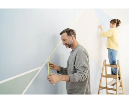 Zwei Personen gestalten eine Wand mit Klebeband für Malerarbeiten und geometrischen Formen.