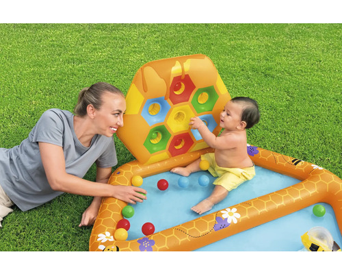 Aufblasbares Planschbecken mit Bienenwabenmuster, Bällen und spielender Mutter mit Baby auf einer Rasenfläche.