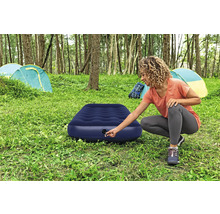 Frau bereitet eine blaue Luftmatratze auf einem Campingplatz mit Zelten im Hintergrund vor.