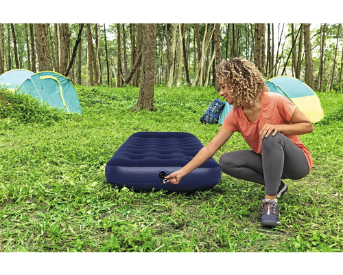 Frau bereitet eine blaue Luftmatratze auf einem Campingplatz mit Zelten im Hintergrund vor.