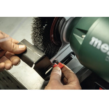 Eine Person verwendet eine Schleifbockmaschine mit Drahtbürste zum Entgraten eines Metallstücks. Metabo Logo.