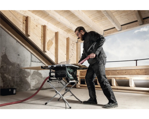 Ein Mann arbeitet mit einer Tischkreissäge in einem Raum mit Holzbalken an der Decke.