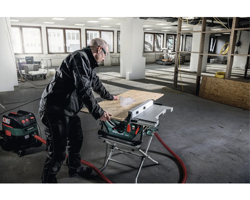 Mann arbeitet mit einer Tischkreissäge und einem Staubsauger in einem Raum.