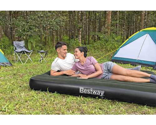 Szene im Wald mit einem Paar, das auf einer Luftmatratze der Marke Bestway liegt, im Hintergrund Zelte und Campingstühle.