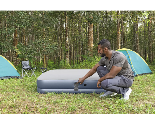 Mann bedient ein aufblasbares Gästebett auf einem Campingplatz mit Zelten und Stühlen.