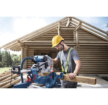 Mann mit Gehörschutz, Schutzbrille und Schutzhelm arbeitet mit einer Kapp- und Gehrungssäge an einem Holzhaus