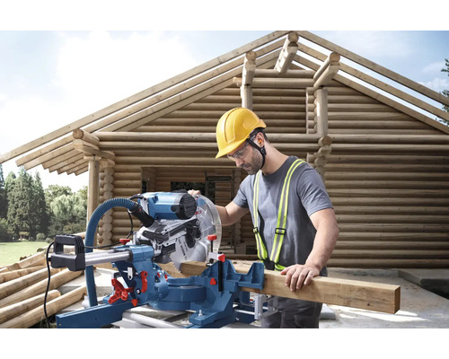 Mann mit Gehörschutz, Schutzbrille und Schutzhelm arbeitet mit einer Kapp- und Gehrungssäge an einem Holzhaus