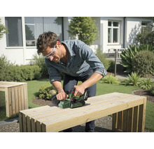 Mann hobelt eine Holzbank mit einem Elektrohobel im Garten.