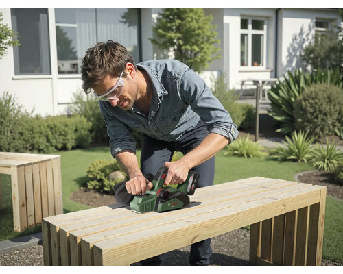 Mann hobelt eine Holzbank mit einem Elektrohobel im Garten.