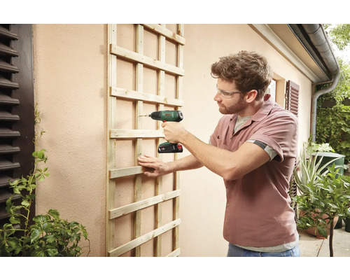 Mann befestigt Holzspalier mit Akkuschrauber an Hauswand