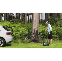 Szene im Freien mit einer Person, die ein Fahrrad mit einem tragbaren Reiniger reinigt. Ein Auto mit Fahrradträger ist ebenfalls zu sehen.