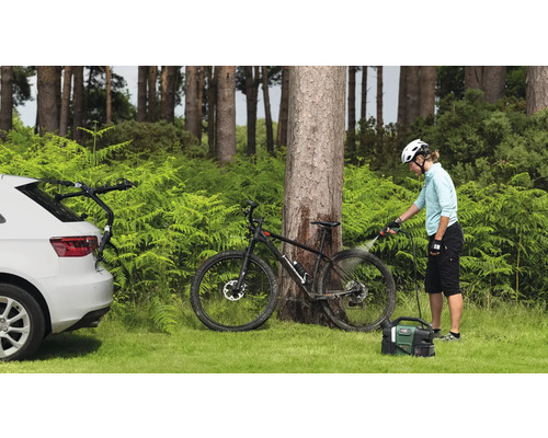 Szene im Freien mit einer Person, die ein Fahrrad mit einem tragbaren Reiniger reinigt. Ein Auto mit Fahrradträger ist ebenfalls zu sehen.