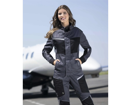 Frau trägt eine graue Arbeitsjacke und Arbeitshose vor einem Flugzeug