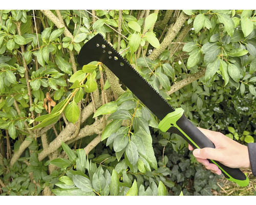 Eine Person hält eine Buschmesser-Gartensäge vor einer grünen Hecke.