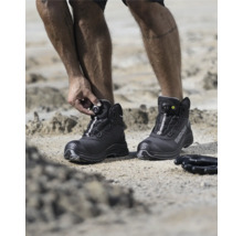 Ein Mann schließt seine schwarzen Ardonsicherheitsstiefel mit Drehradverschluss auf einem sandigen Untergrund.
