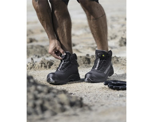 Ein Mann schließt seine schwarzen Ardonsicherheitsstiefel mit Drehradverschluss auf einem sandigen Untergrund.
