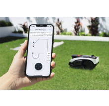 Person hält Smartphone mit App zur Steuerung eines Rasenmähroboters in der Hand. Im Hintergrund befindet sich der Rasenmähroboter auf einer Rasenfläche.
