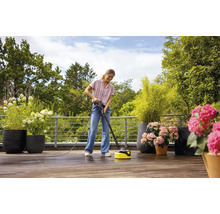 Frau reinigt Holzterrasse mit einem Terrassenreiniger unter freiem Himmel, umgeben von Topfpflanzen.