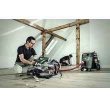 Zwei Handwerker arbeiten in einem hellen Raum mit Holzbalken. Einer bedient eine Metabo Kapp- und Gehrungssäge, während der andere Arbeiten am Boden ausführt. Ein Metabo Staubsauger ist angeschlossen.
