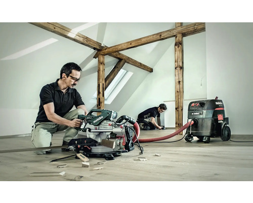 Zwei Handwerker arbeiten in einem hellen Raum mit Holzbalken. Einer bedient eine Metabo Kapp- und Gehrungssäge, während der andere Arbeiten am Boden ausführt. Ein Metabo Staubsauger ist angeschlossen.