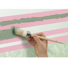 Eine Person streicht mit einem Rundpinsel eine grüne Linie auf eine Wand mit rosa und weißen Streifen